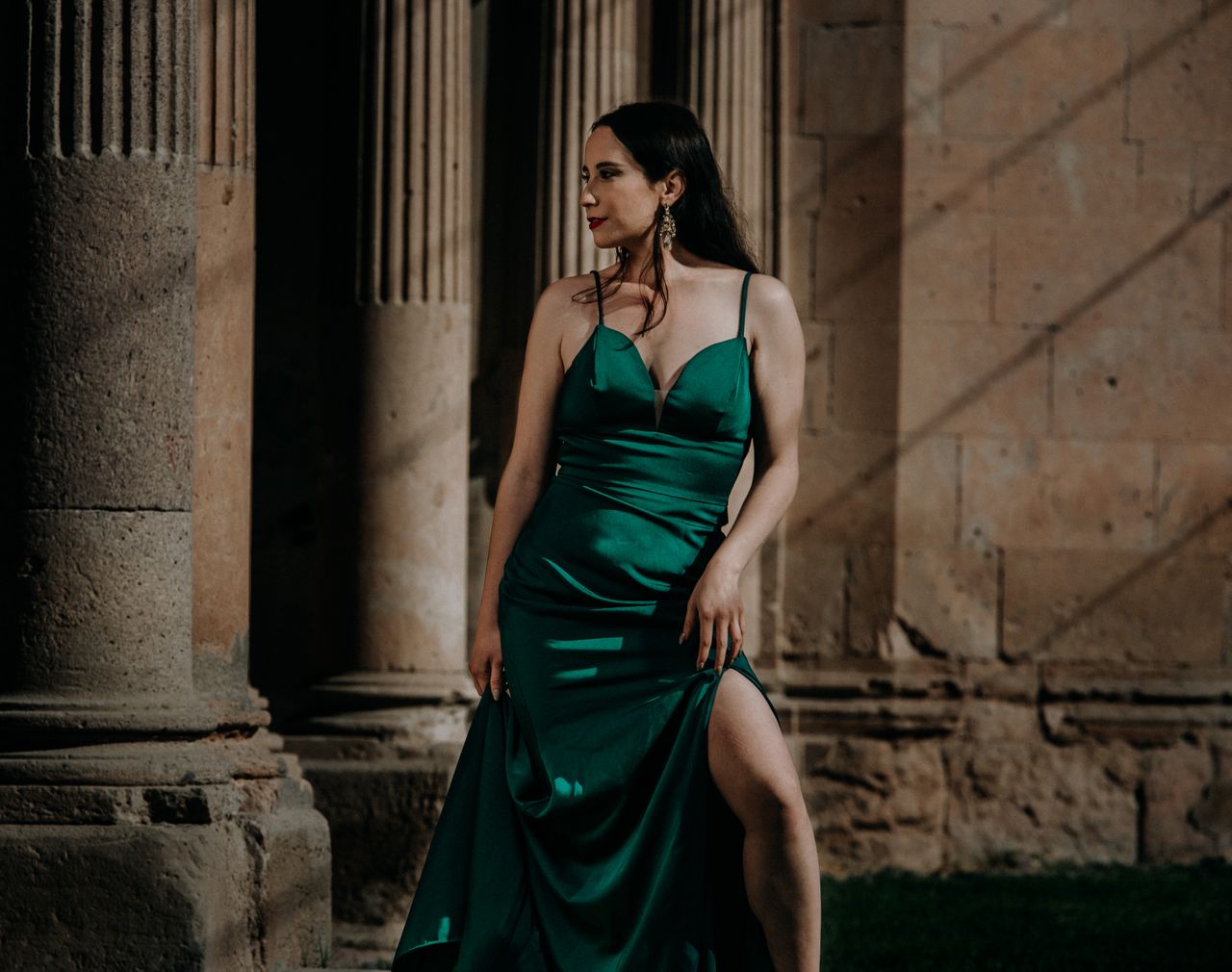 Services Model in a green dress standing elegantly by ancient stone columns, exuding sophistication.