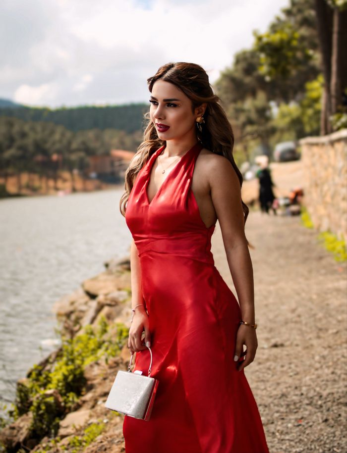 Services A stylish woman in a red maxi dress posing elegantly by the river on a sunny day.