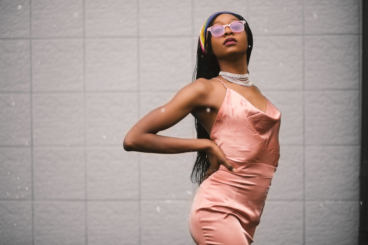 About Elegant young woman in a pink dress and sunglasses posing with confidence against a neutral background.